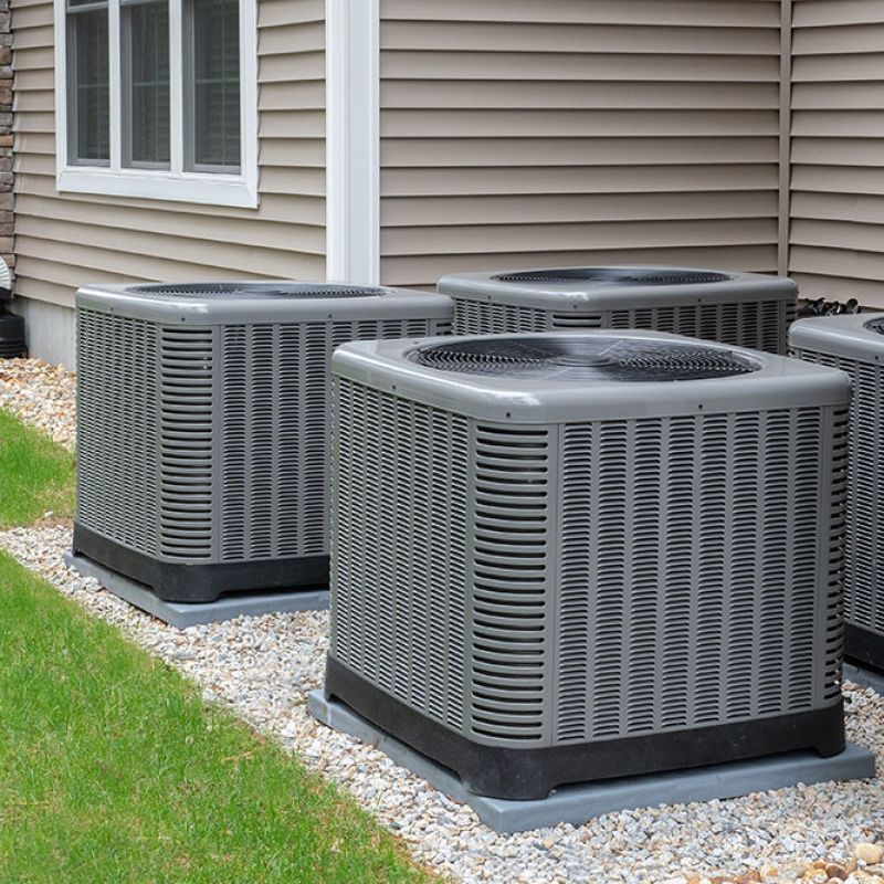 Outdoor HVAC units beside a house, with siding and stone exterior, surrounded by green grass and gravel.