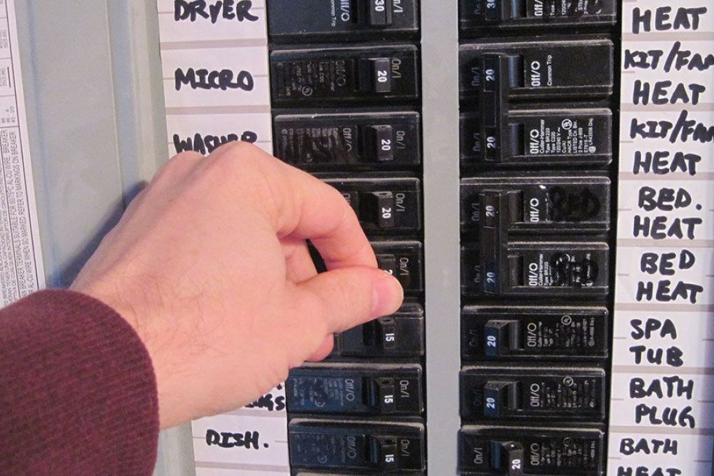 Hand switches off circuit breaker in labeled electrical panel.