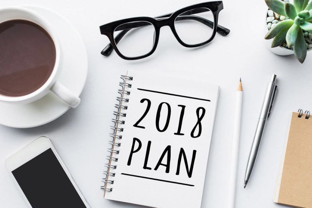 Desk essentials for planning: glasses, coffee, smartphone, and a 2018 planner with stationery on a white surface.