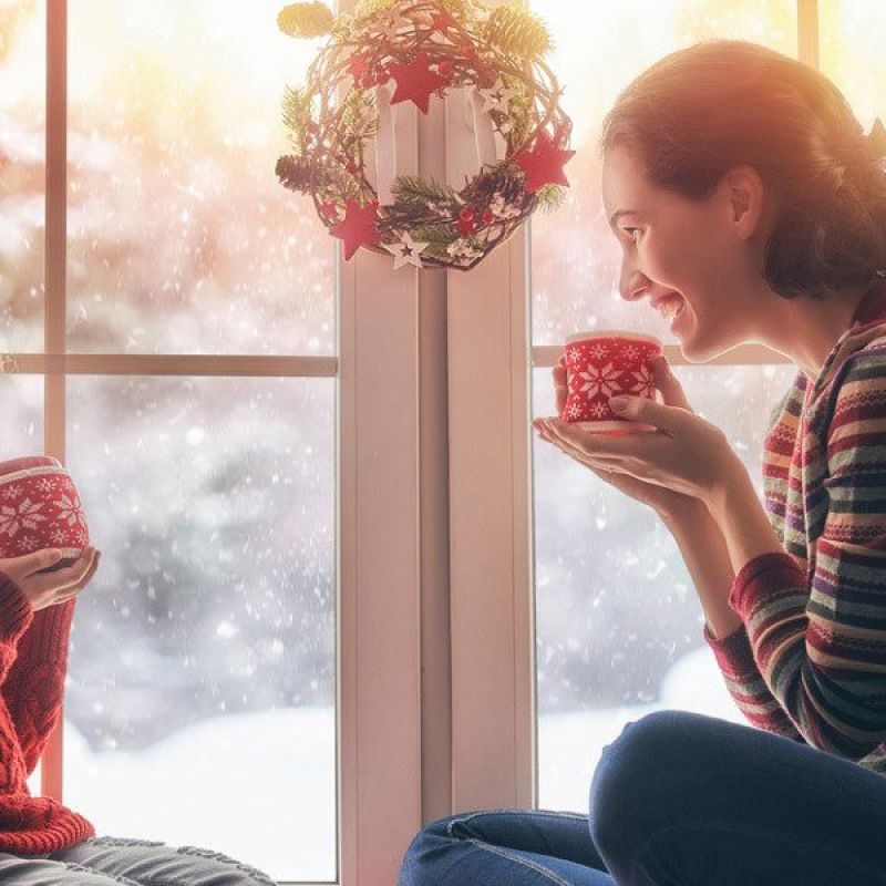 Mother and daughter enjoying hot cocoa by a snowy window, festive wreath in background. Cozy winter scene.