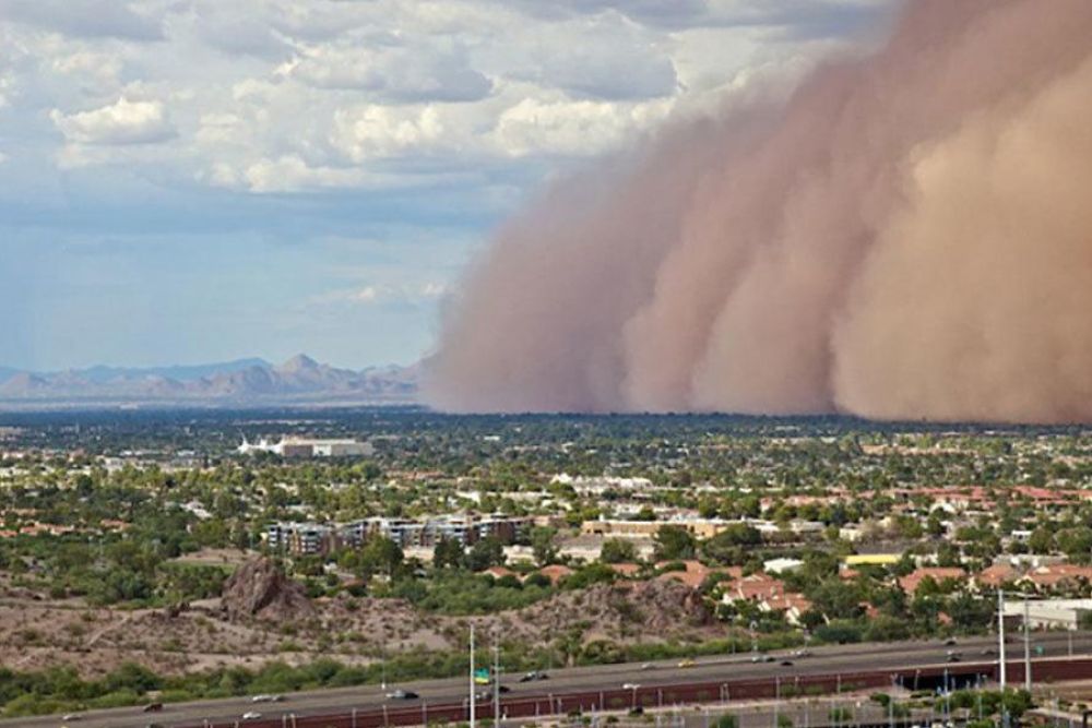 Massive dust storm (haboob) sweeping across a desert city under cloudy skies, with mountains in the background.