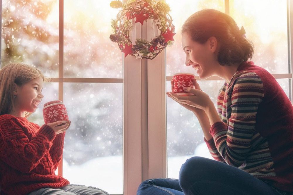 Mother and daughter enjoying hot cocoa by a snowy window, festive wreath in background. Cozy winter scene.