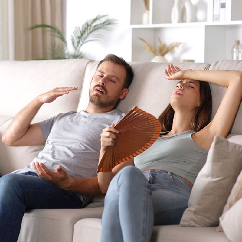 Couple on sofa feeling hot and tired, woman fanning for relief. Home summer heat and cooling concept.