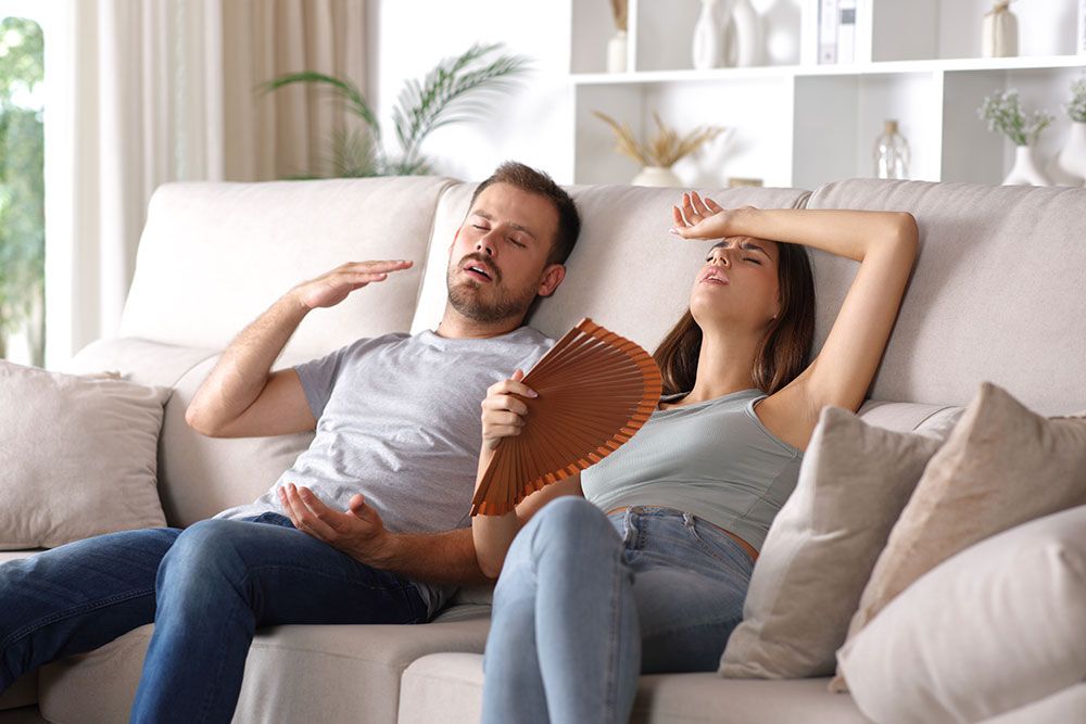 Couple on sofa feeling hot and tired, woman fanning for relief. Home summer heat and cooling concept.