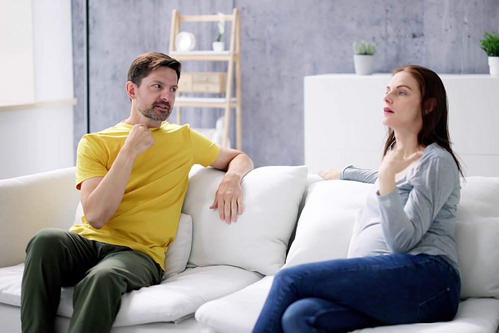 Pregnant woman and man communicating in sign language on a white sofa in a modern living room.