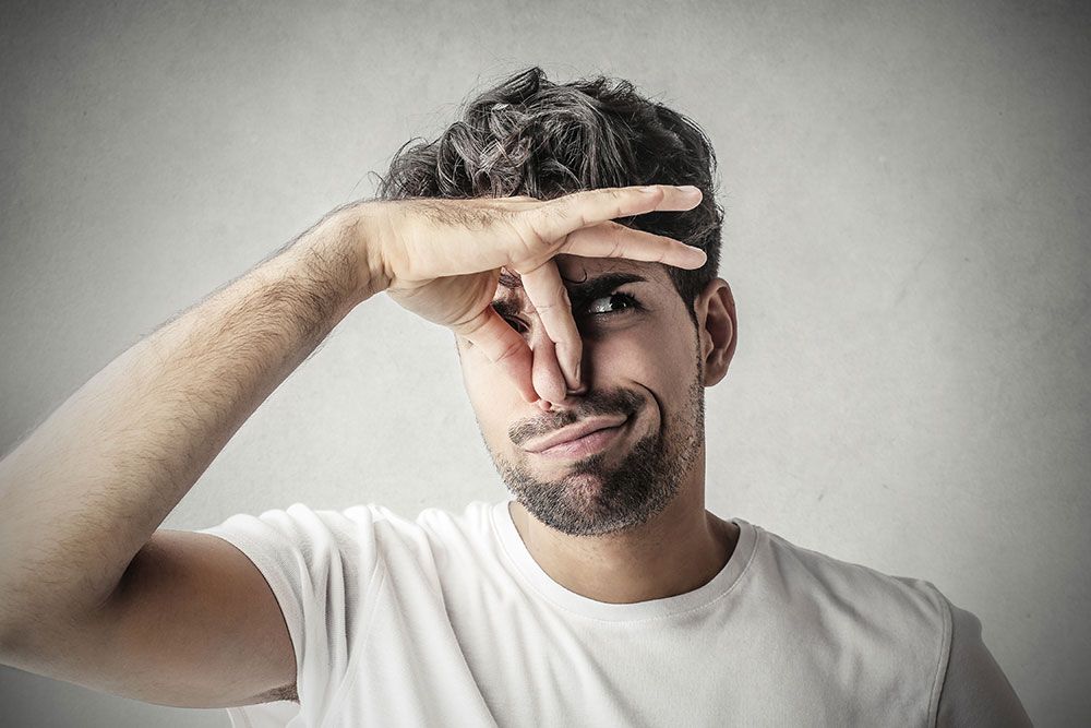 Man in white shirt pinching nose, expressing disgust or bad smell against gray background.
