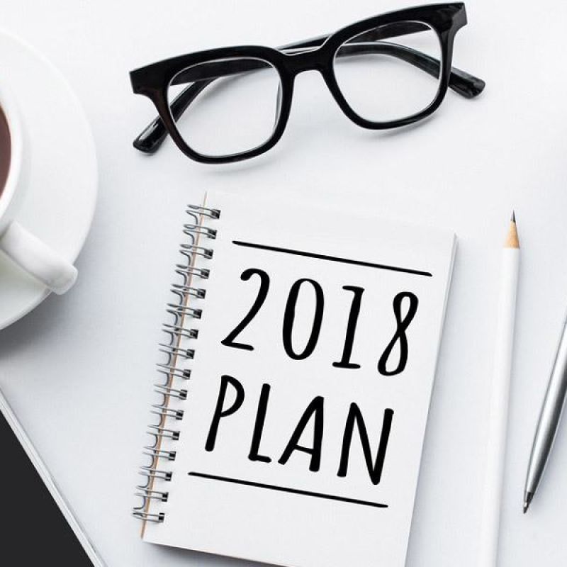 Desk essentials for planning: glasses, coffee, smartphone, and a 2018 planner with stationery on a white surface.