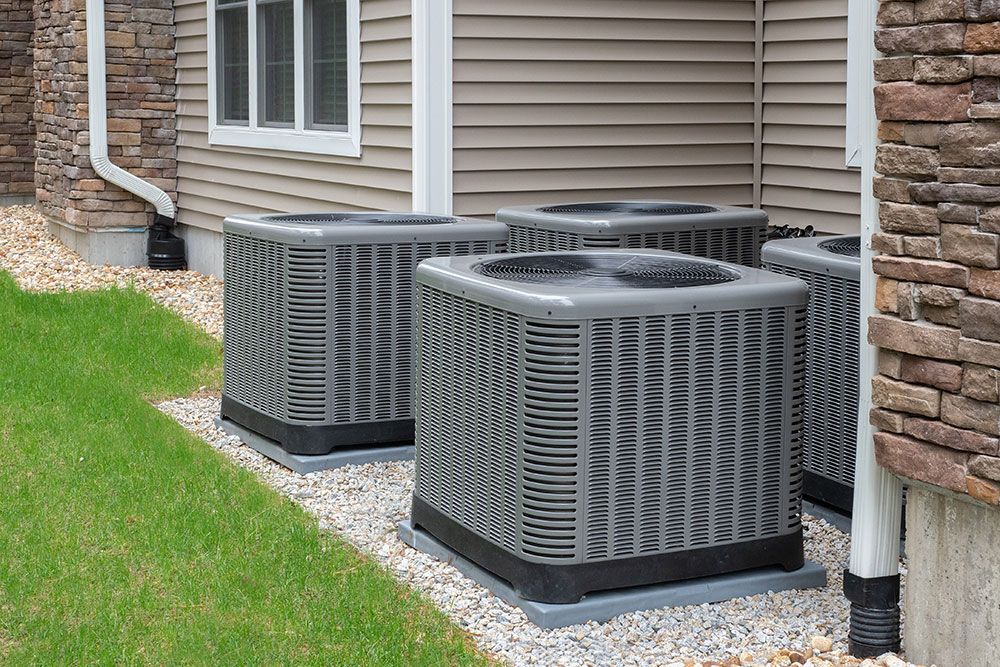 Outdoor HVAC units beside a house, with siding and stone exterior, surrounded by green grass and gravel.