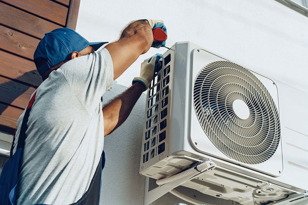 HVAC technician in a blue cap and gloves secures an outdoor air conditioner condenser to a building wall with a screwdriver.",