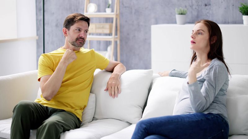 Pregnant woman and man communicating in sign language on a white sofa in a modern living room.