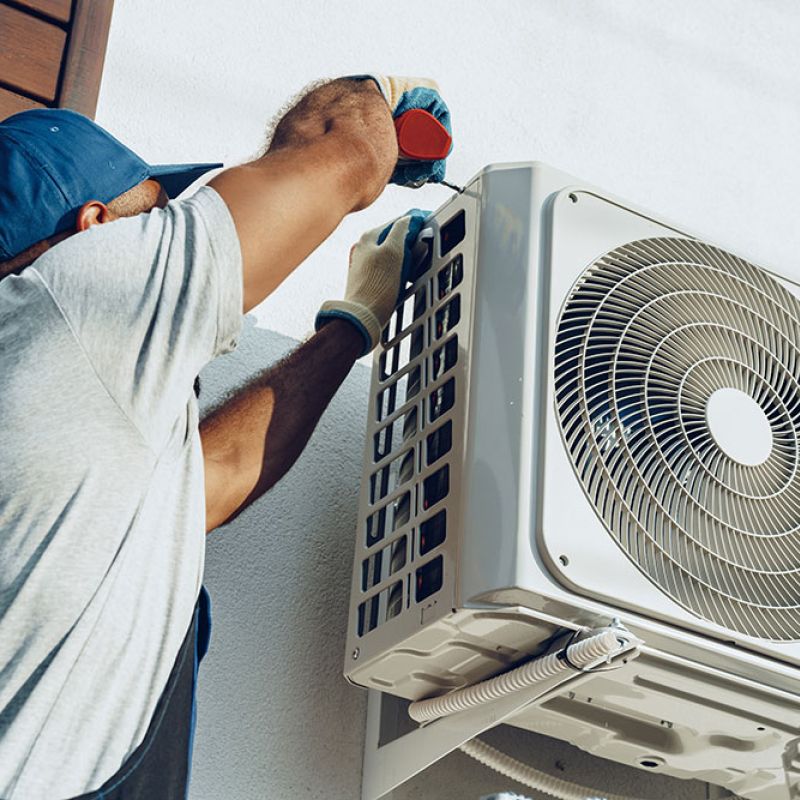 HVAC technician in a blue cap and gloves secures an outdoor air conditioner condenser to a building wall with a screwdriver.",