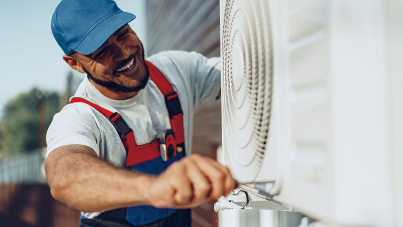 Smiling technician in blue cap repairs outdoor air conditioning unit with a screwdriver.