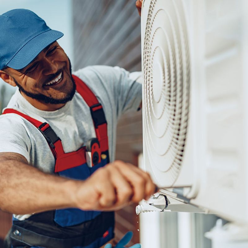 Smiling technician in blue cap repairs outdoor air conditioning unit with a screwdriver.