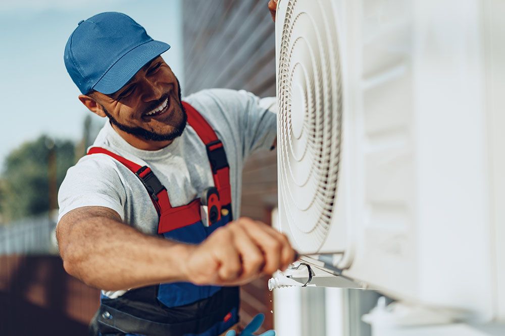 Smiling technician in blue cap repairs outdoor air conditioning unit with a screwdriver.