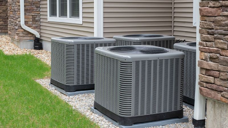 Outdoor HVAC units beside a house, with siding and stone exterior, surrounded by green grass and gravel.